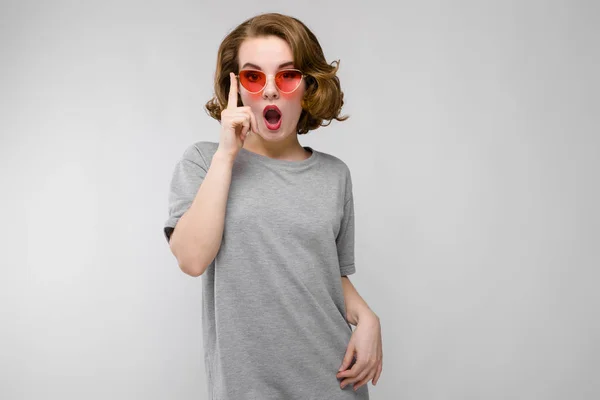 Encantadora joven con una camiseta gris sobre un fondo gris. Chica feliz en gafas rojas — Foto de Stock