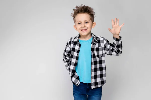 Um menino bonito em uma camisa xadrez, camisa azul e jeans fica em um fundo cinza. O menino sorri e mostra a palma da mão — Fotografia de Stock