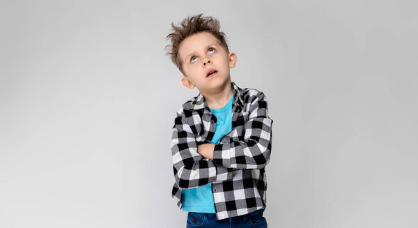 A handsome boy in a plaid shirt, blue shirt and jeans stands on a gray background. The boy folded his arms over his chest. The boy has closed his eyes