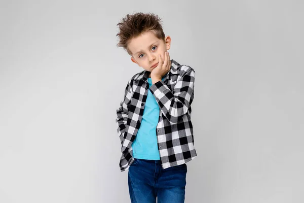 Красивый мальчик в клетчатой рубашке, голубой рубашке и джинсах стоит на сером фоне. Мальчик поддерживает подбородок рукой. — стоковое фото