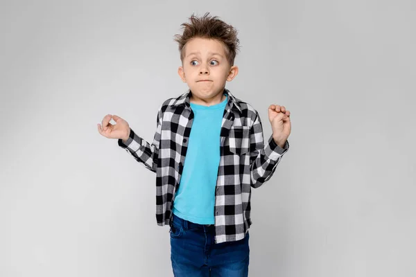 Yakışıklı bir çocuk bir ekose gömlek, mavi tişört ve kot gri bir arka plan üzerinde duruyor. Çocuk elleri her iki yönde yaymak — Stok fotoğraf