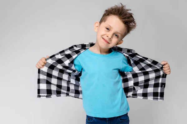 Um menino bonito em uma camisa xadrez, camisa azul e jeans fica em um fundo cinza. O rapaz puxa a camisa para trás — Fotografia de Stock