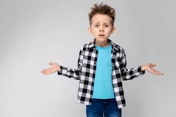 Um menino bonito em uma camisa xadrez, camisa azul e jeans fica em um fundo cinza. O menino estendeu as mãos em ambas as direções — Fotografia de Stock