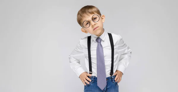 Un niño encantador con una camisa blanca, tirantes, corbata y pantalones vaqueros ligeros se levanta sobre un fondo gris. el niño con gafas inclinó la cabeza a su lado —  Fotos de Stock