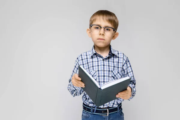 一个迷人的男孩与镶有衬衫和轻牛仔裤站在灰色的背景。那男孩手里拿着一本书。戴眼镜的男孩 — 图库照片
