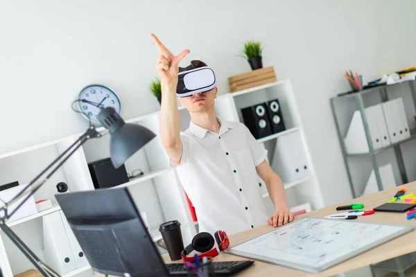 Um jovem de óculos de realidade virtual fica perto da mesa e puxa o braço para cima. Um jovem polegar uma página virtual . — Fotografia de Stock