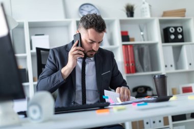 Bir adam masada, ofisinde oturuyor telefonda konuşurken ve belgeler üzerinden çevirme.