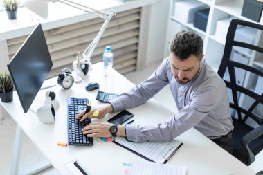 Ofiste genç bir adam bir masada oturuyor, bir işaretleyici elinde tutuyor ve belgeler ve bir bilgisayar ile çalışır.