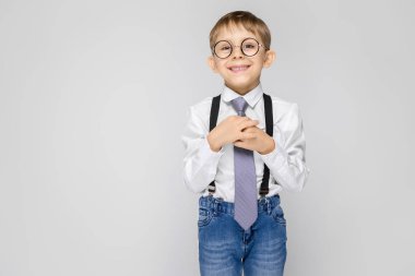 Beyaz bir gömlek, pantolon askısı, kravat ve ışık kot büyüleyici bir çocuk gri bir arka plan üzerinde duruyor. Oğlan gülümsüyor ve el kravatını tutar