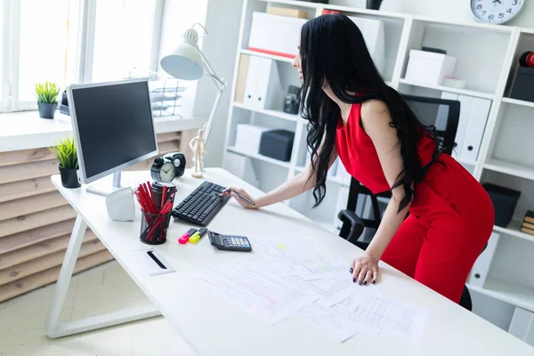 Een mooi jong meisje staat in de buurt van een bureau. Het meisje werkt met documenten, rekenmachine en computer. — Stockfoto