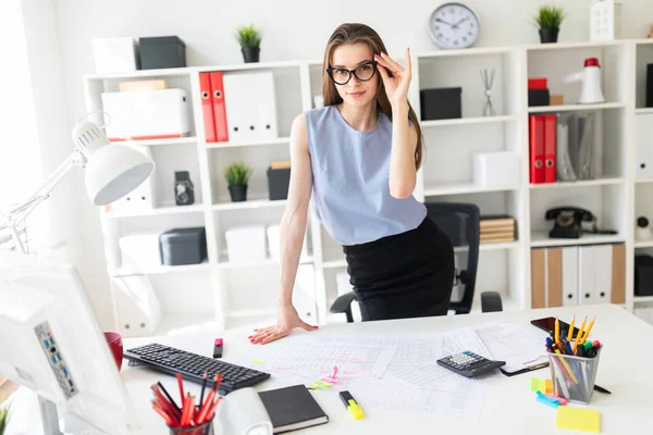 Hermosa joven en la oficina está de pie cerca de la mesa y fija sus gafas con la mano . — Foto de Stock