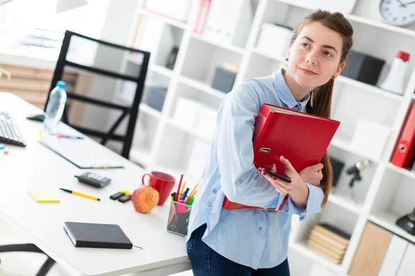 En ung flicka på kontoret är stående, lutad mot ett bord, och håller en telefon och en mapp med dokument. — Stockfoto