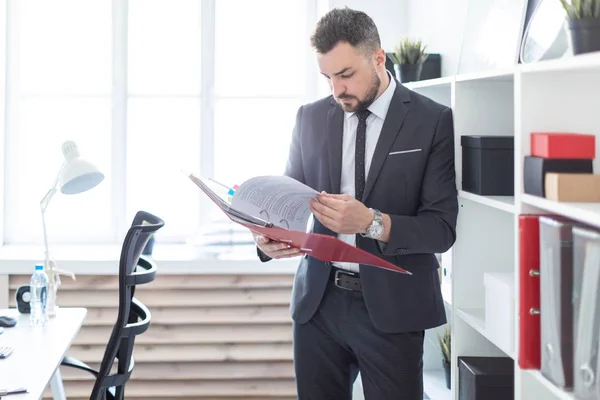 The man is standing near the rack in the office and scrolls through the folder with the documents. — Stock Photo, Image