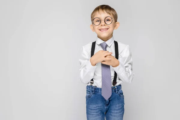 Un niño encantador con una camisa blanca, tirantes, corbata y pantalones vaqueros ligeros se levanta sobre un fondo gris. Un niño sonríe y sostiene su corbata de mano — Foto de Stock