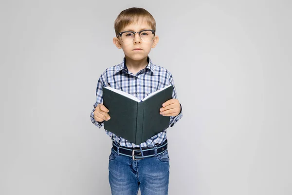 Vor grauem Hintergrund steht ein charmanter Junge mit eingelegtem Hemd und heller Jeans. Der Junge hält ein Buch in der Hand. Junge mit Brille — Stockfoto