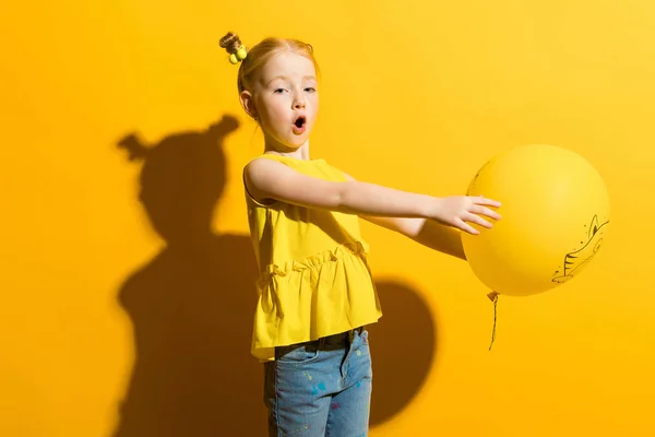 Ragazza con i capelli rossi su sfondo giallo. La ragazza tiene un palloncino giallo sulle braccia tese . — Foto Stock