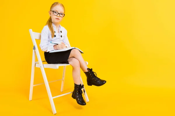 Chica con el pelo rojo sobre un fondo amarillo. Una chica encantadora en gafas transparentes se sienta en una silla blanca y toma notas en un cuaderno . — Foto de Stock