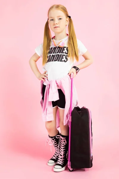 Κορίτσι με κόκκινο ουρές σε ροζ φόντο. Ένα γοητευτικό κορίτσι με έντονο σπορ ρούχα κρατώντας μια βαλίτσα από τη λαβή. — Φωτογραφία Αρχείου