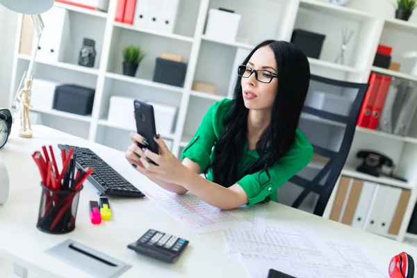 Mladá dívka v brýlích sedí v kanceláři na stole a drží telefon v ruce. — Stock fotografie