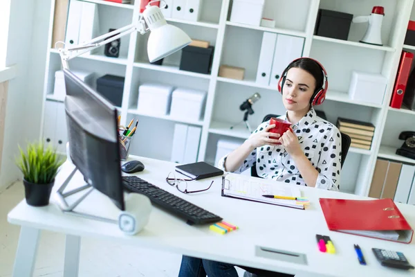 Een jong meisje zit in de koptelefoon aan een tafel in het kantoor, houdt een rode kop in haar handen en kijkt naar de monitor. — Stockfoto