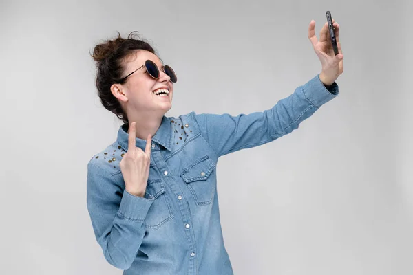Μελαχρινή κοπέλα με τα στρογγυλά γυαλιά. Τρίχες που συγκεντρώθηκαν σε ένα κουλούρι. Κορίτσι με ένα μαύρο τηλέφωνο. Το κορίτσι κάνει selfie. — Φωτογραφία Αρχείου