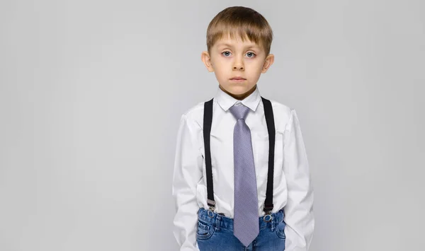 Ein charmanter Junge in weißem Hemd, Hosenträgern, Krawatte und heller Jeans steht vor grauem Hintergrund — Stockfoto