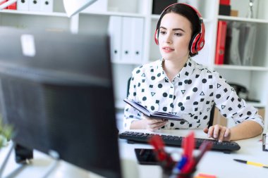 Güzel genç kız kulaklık Office masada oturur, onun elinde bir defter tutan ve klavyede yazdırır. Alan, kız odak derinliği ile fotoğraf.