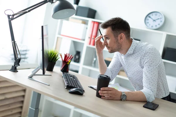 Молодой человек стоит возле стола в офисе, держа в руках карандаш и стакан кофе. Молодой человек работает с документами и компьютером . — стоковое фото