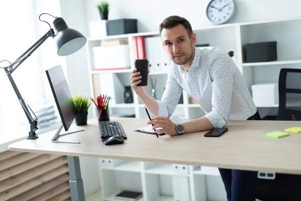 Молодой человек стоит возле стола в офисе, держа в руках карандаш и стакан кофе. Молодой человек работает с документами и компьютером . — стоковое фото