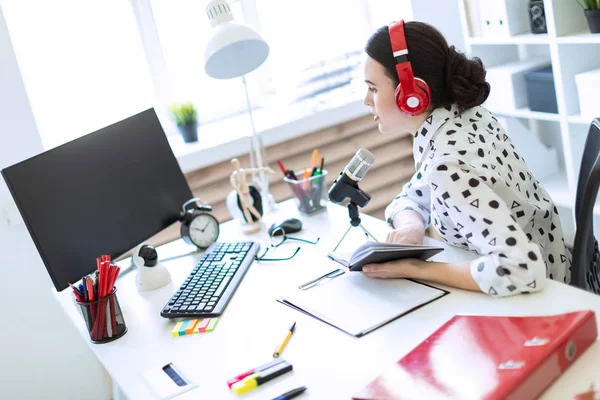 Menina bonita senta-se em fones de ouvido e com um bloco de notas na mesa no escritório e fala no microfone . — Fotografia de Stock