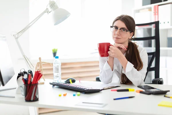 En ung flicka sitter vid ett bord på kontoret och innehar en röd kopp i händerna. — Stockfoto