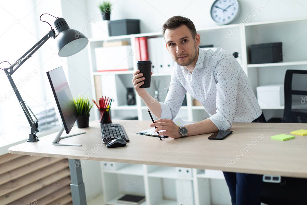 A young man is standing near a table in the office, holding a pencil and a glass of coffee. A young man works with documents and a computer.