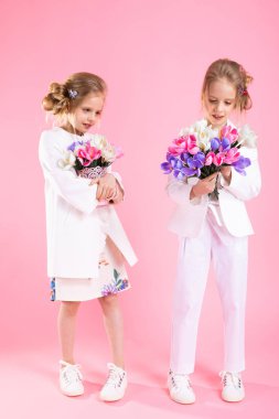 İkiz kızlar çiçek demetleri ile hafif giysiler içinde pembe bir arka plan üzerinde durmak.