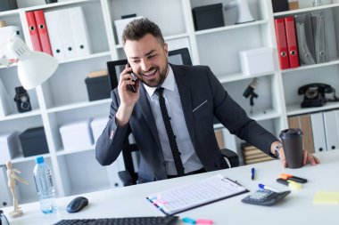 Bir adam ofiste masa başında oturan, telefonda konuşurken ve bir bardak kahve elinde tutan.
