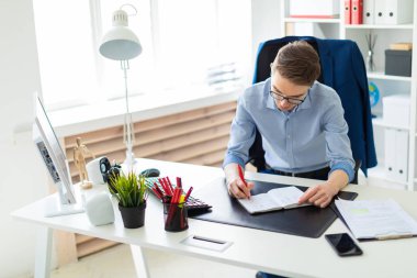 Genç bir adam bir bilgisayar masası ofisinde oturuyor ve bir not defterinde yazıyor.