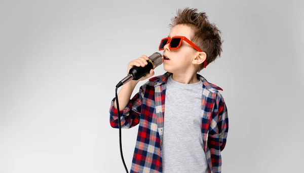 Um menino bonito em uma camisa xadrez, camisa cinza e jeans fica em um fundo cinza. Um rapaz a usar óculos de sol. Rapaz ruivo canta no microfone — Fotografia de Stock