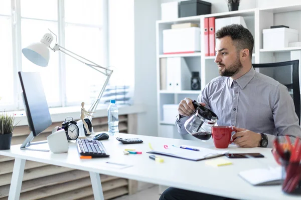 Un joven en la oficina se sienta en una mesa, mira el monitor y vierte café en una taza . — Foto de Stock