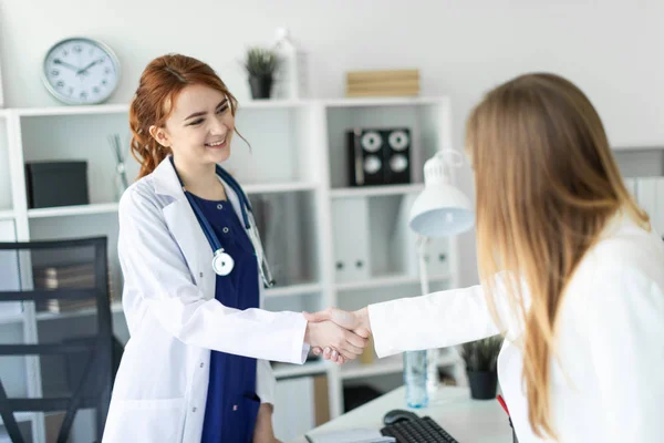 Uma menina bonita com um casaco branco está de pé perto da mesa no escritório e apertando as mãos com o paciente. O paciente agradece ao médico . — Fotografia de Stock