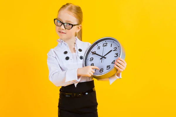 Mädchen mit roten Haaren auf gelbem Hintergrund. ein charmantes Mädchen mit durchsichtiger Brille hält eine Uhr. — Stockfoto