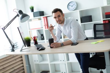 Genç bir adam bir bardak kahve ve bir kalem tutan bir tablo ofiste, yakın duruyor. Genç bir adam belgeleri ve bir bilgisayar ile çalışır.