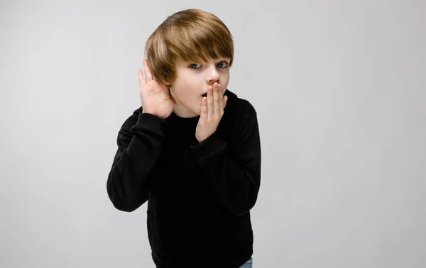 Charmiga tonåring med blont hår och mörka ögon. Pojken stänger munnen med ena handen, ger sin andra hand till sitt öra — Stockfoto