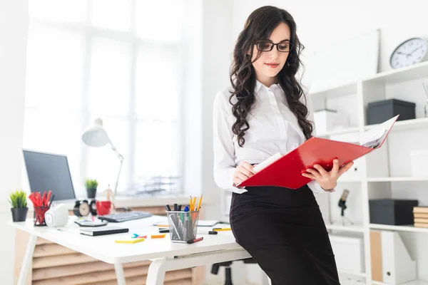 Vacker ung flicka står i office, lutande på skrivbord och bläddrar genom röda mapp med dokument. — Stockfoto