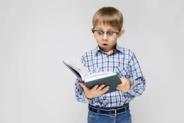 Vor grauem Hintergrund steht ein charmanter Junge mit eingelegtem Hemd und heller Jeans. Der Junge hält ein Buch in der Hand. Junge mit Brille — Stockfoto