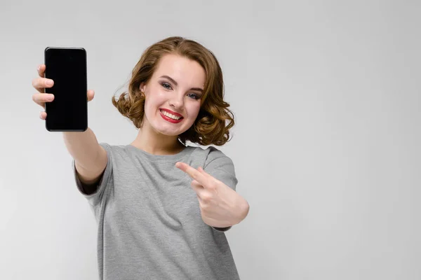 Menina encantadora em uma camiseta cinza em um fundo cinza. A menina está segurando um telefone — Fotografia de Stock