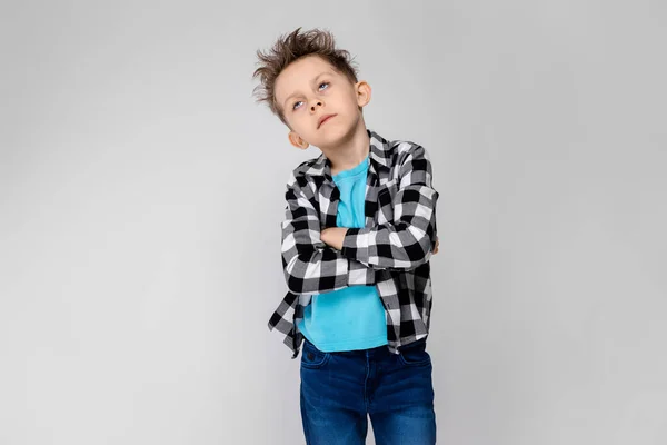 Un chico guapo con camisa a cuadros, camisa azul y pantalones vaqueros está parado sobre un fondo gris. El niño dobló los brazos sobre su pecho. El chico ha cerrado los ojos. —  Fotos de Stock