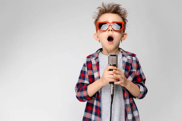 Een knappe jongen in een plaid shirt, grijs shirt en spijkerbroek staat op een grijze achtergrond. Een jongen dragen van een zonnebril. Roodharige jongen zingt in de microfoon — Stockfoto