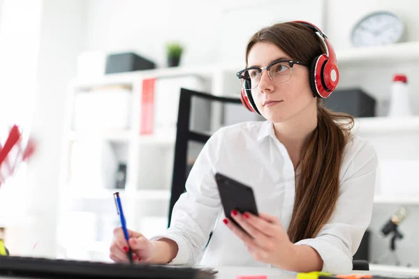 Une jeune fille est assise dans un casque à une table dans le bureau et tient un stylo et un téléphone dans sa main . — Photo