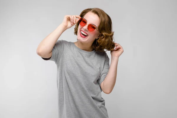 Menina encantadora em uma camiseta cinza em um fundo cinza. Menina feliz em óculos vermelhos. A menina ri — Fotografia de Stock