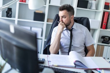 Bir adam ofiste bir sandalyede oturur ve bir kalem elinde tutuyor. Sandalyenin arkasında ceketini asmak.