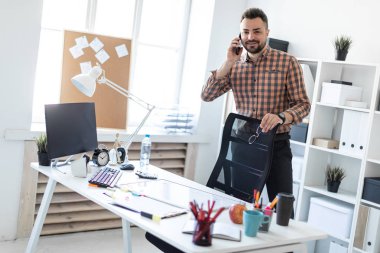 Bir adam bilgisayar masası yakınındaki ofiste standları, elini koltuğun arkasına koyar ve telefonda konuşuyor.